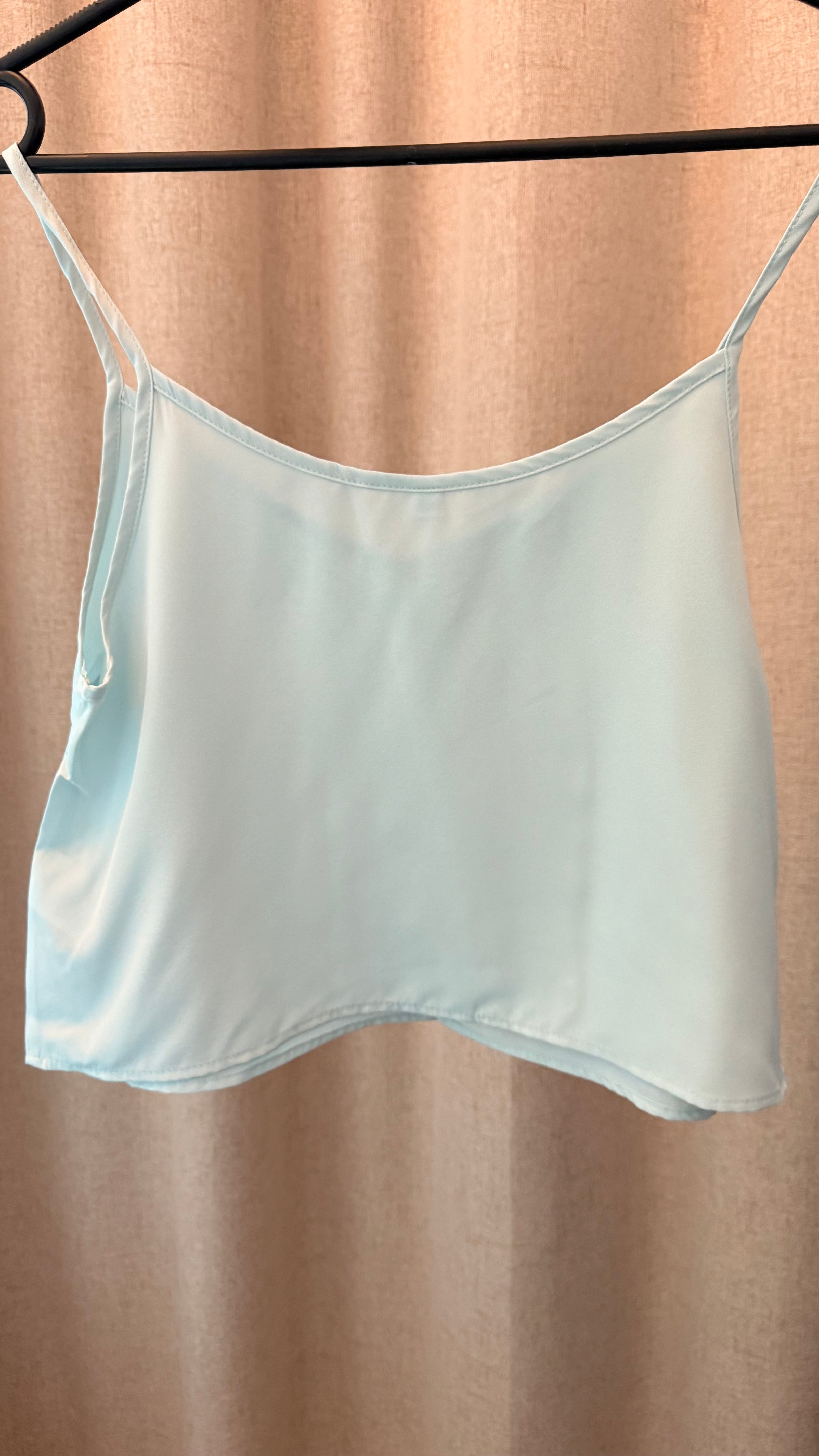 Light blue satin camisole on a hanger against a beige curtain background