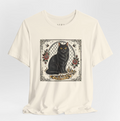 An off-white short-sleeve t-shirt with a central graphic print of a black cat sitting in front of a chain-link fence, surrounded by red roses and birds, on a light gray background.