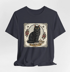 A dark navy blue short-sleeve t-shirt with a square graphic print on the chest featuring a black cat, red roses, and traditional tattoo-style details on a cream background.