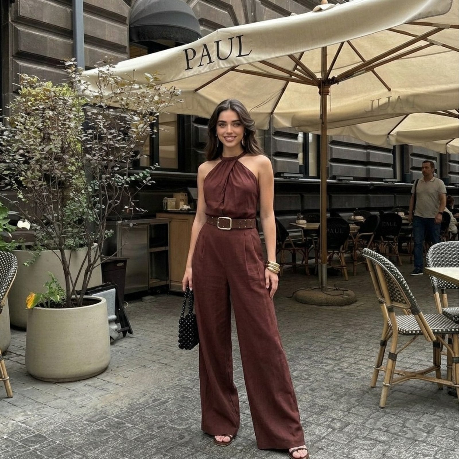 Woman in a brown jumpsuit standing under a large umbrella with 'PAUL' branding in an outdoor setting.