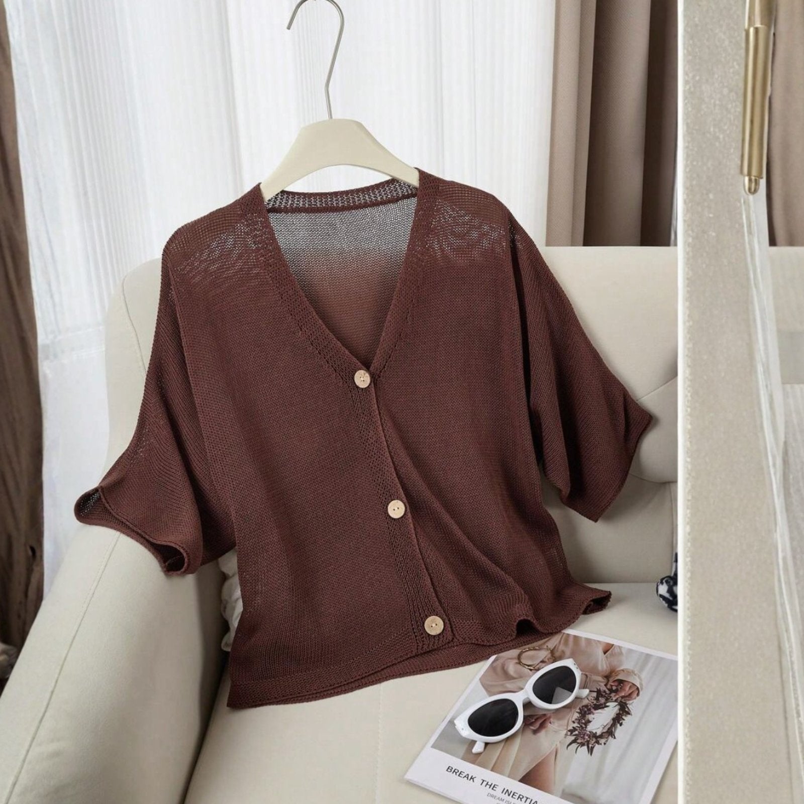 Brown cardigan on a hanger with sunglasses and magazine on a white couch