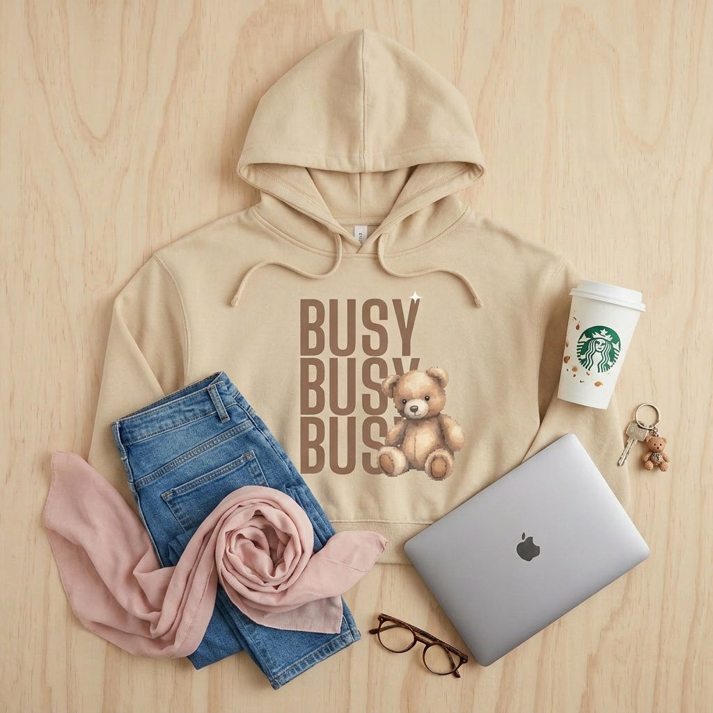 Beige hoodie with 'BUSY BUSY BUSY' text and teddy bear graphic, paired with jeans, a pink scarf, laptop, Starbucks cup, and glasses on a wooden surface.
