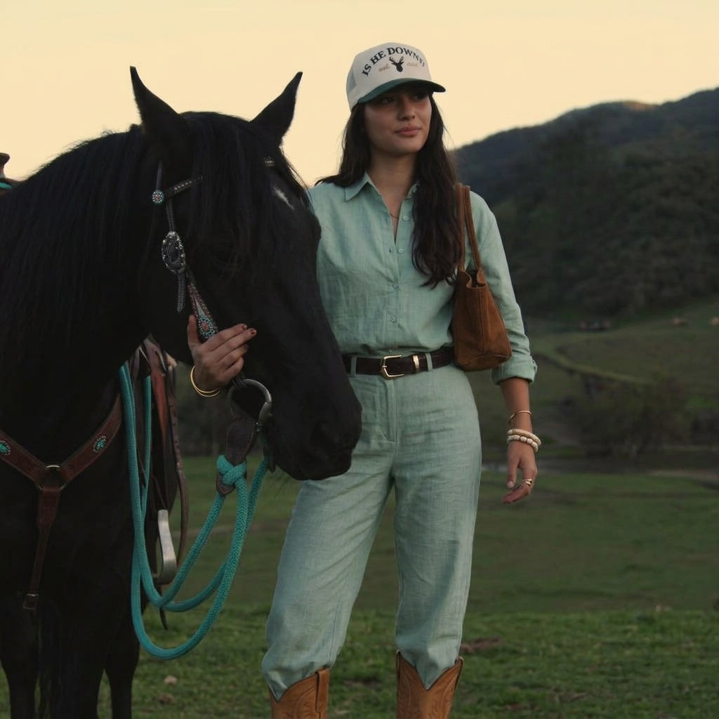 A woman stands outdoors in a grassy field next to a black horse, wearing a light sage green linen shirt and matching pants with brown boots, a brown shoulder bag, and a white baseball cap.