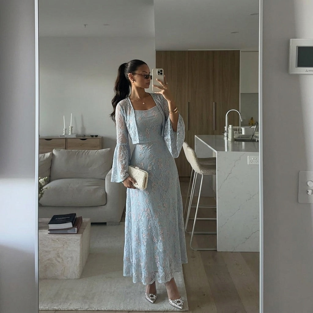 A woman takes a mirror selfie in a modern living room, wearing a light blue floral lace maxi dress with a matching cropped jacket and holding a small cream clutch.