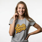 A smiling young woman with long light brown hair wears a heather gray t-shirt featuring a retro-style yellow and purple graphic that reads 'Smile, please'. She is posing against a clean, light gray studio background.