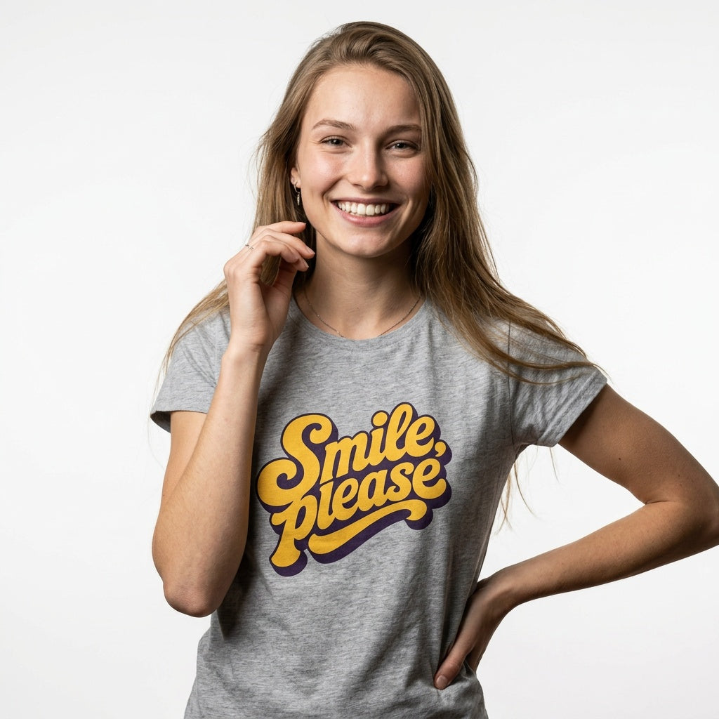A smiling young woman with long light brown hair wears a heather gray t-shirt featuring a retro-style yellow and purple graphic that reads 'Smile, please'. She is posing against a clean, light gray studio background.