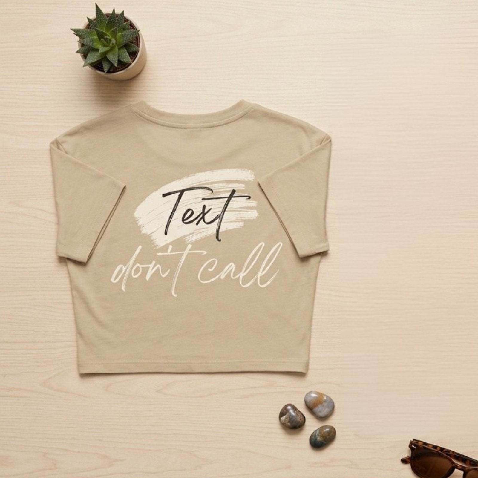 Beige t-shirt with 'Text don't call' text on a light wooden surface with decorative items.