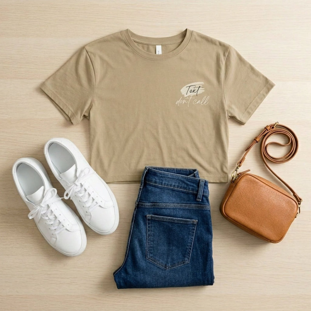 Beige t-shirt with text, white sneakers, blue jeans, and a brown crossbody bag on a beige surface.