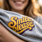 A close-up of a person wearing a heather gray t-shirt with a retro-style graphic that reads 'Smile, please' in yellow and purple.