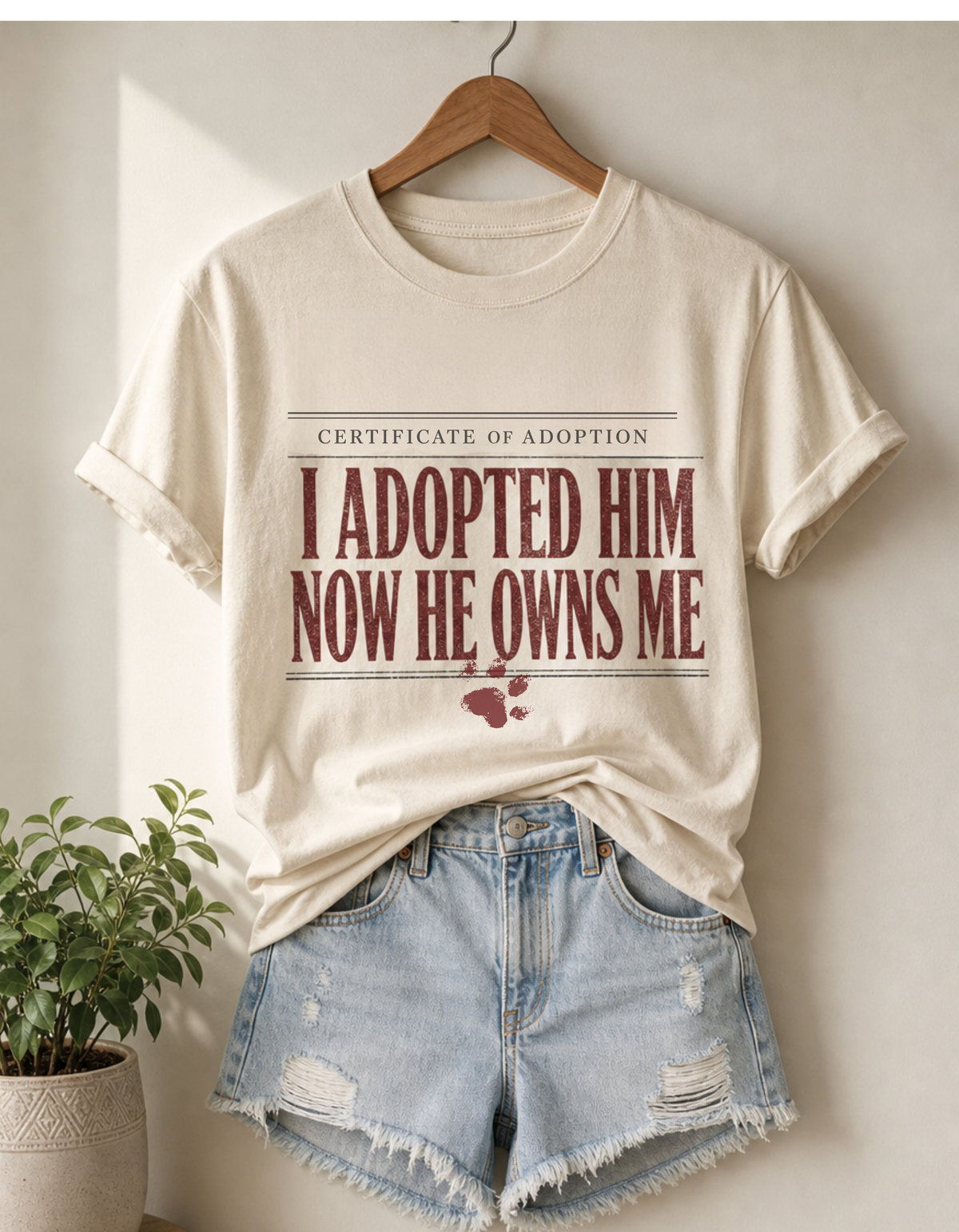 A cream-colored graphic t-shirt on a wooden hanger, styled with light-wash distressed denim shorts. The t-shirt features a 'Certificate of Adoption' style graphic with the text 'I ADOPTED HIM NOW HE OWNS ME' in dark red lettering and a small paw print.