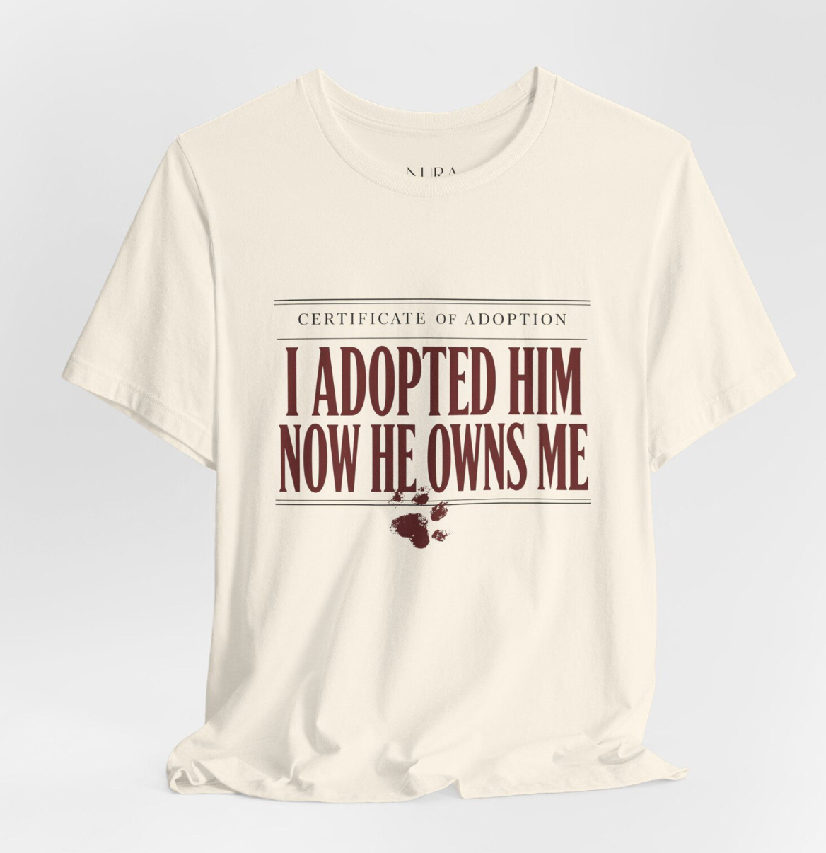 An off-white short-sleeve t-shirt with a graphic print on the front. The print features the text 'CERTIFICATE OF ADOPTION' in a thin serif font, followed by 'I ADOPTED HIM NOW HE OWNS ME' in large, bold, dark red letters.
