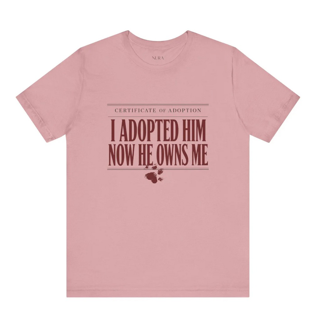 A dusty pink crewneck t-shirt with a graphic print on the front that reads 'CERTIFICATE OF ADOPTION I ADOPTED HIM NOW HE OWNS ME' in dark red text, accompanied by a small paw print and heart graphic below.