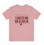 A dusty pink crewneck t-shirt with a graphic print on the front that reads 'CERTIFICATE OF ADOPTION I ADOPTED HIM NOW HE OWNS ME' in dark red text, accompanied by a small paw print and heart graphic below.
