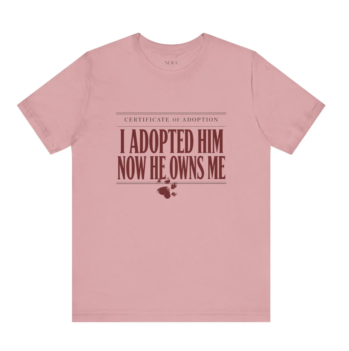 A dusty pink crewneck t-shirt with a graphic print on the front that reads 'CERTIFICATE OF ADOPTION I ADOPTED HIM NOW HE OWNS ME' in dark red text, accompanied by a small paw print and heart graphic below.