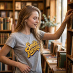 A young woman with long blonde hair stands in a library or bookstore, wearing a heather gray t-shirt with a yellow and purple retro-style graphic that reads 'Smile, please'. She is reaching for a book on a wooden shelf next to her.